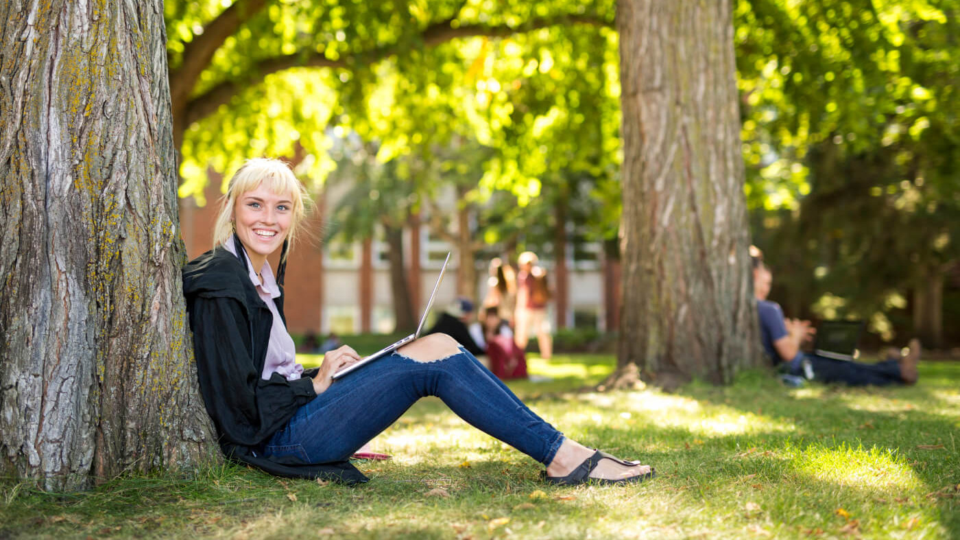 Student sits by a tree on a laptop