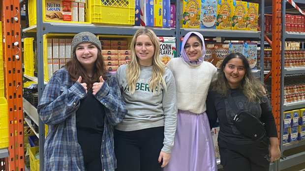 Four students pose inside the Yukon Food Bank building by shelves stocked with nonperishable foods