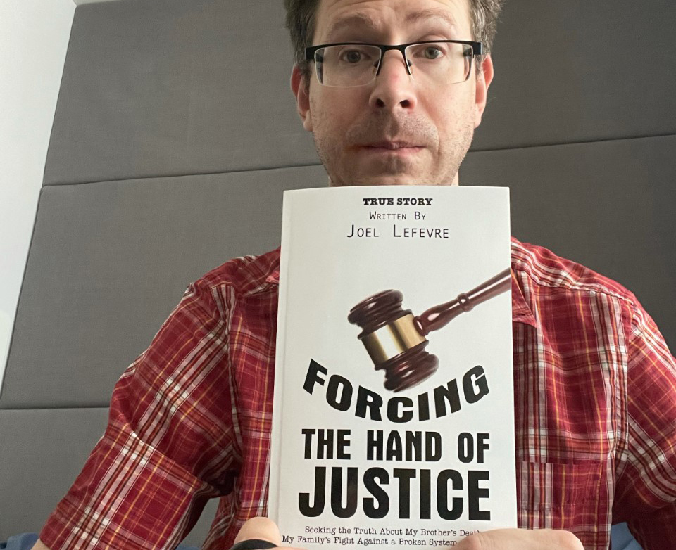 Joel Lefèvre, wearing glasses, holding up his book, Forcing the Hand of Justice.