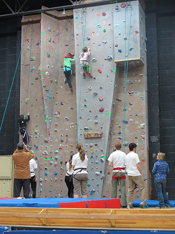 A photo of 2 people on the climbing wall, with a number of people on the floor below.