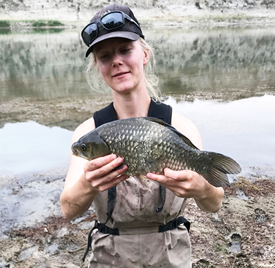 MSc student Cait Donadt stands on river bank holding a very large Prussian carp that's approximately 30 centimetres long.