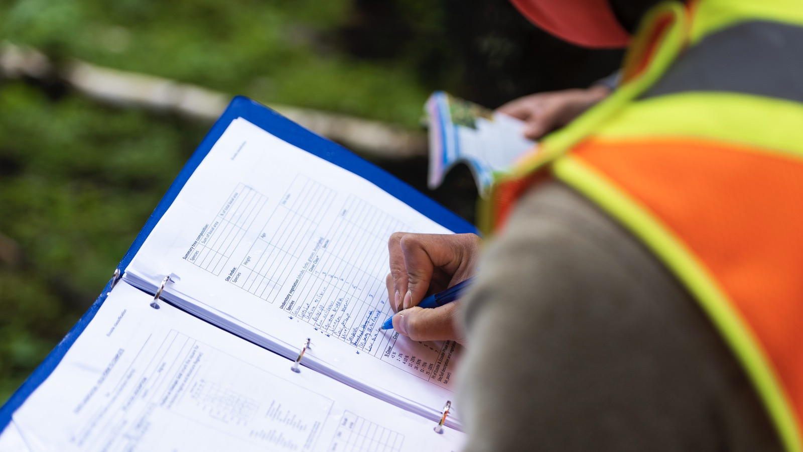 Over-the-shoulder image of a researcher making notes in a binder. Researcher is wearing a safety vest and appears to be in a grassy field.