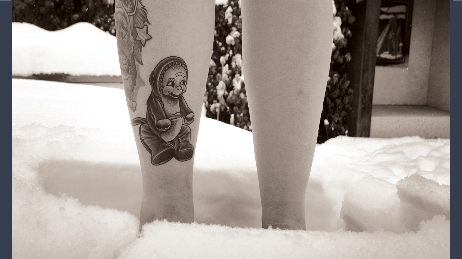 A black and white photograph of bare legs standing in deep snow, showcasing a fresh tattoo of a 1950’s stuffed monkey on the person's right shin.