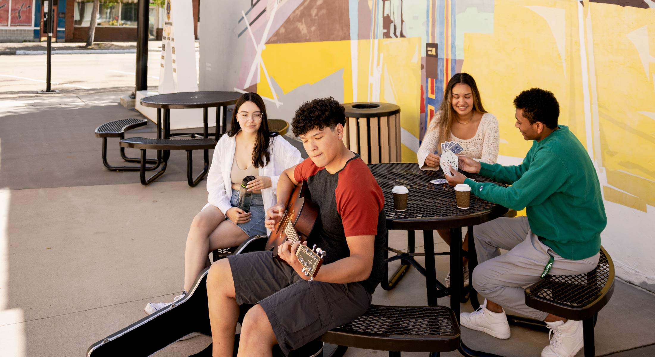 Students sitting around a table outside on campus, playing cards, guitar and drinking coffee