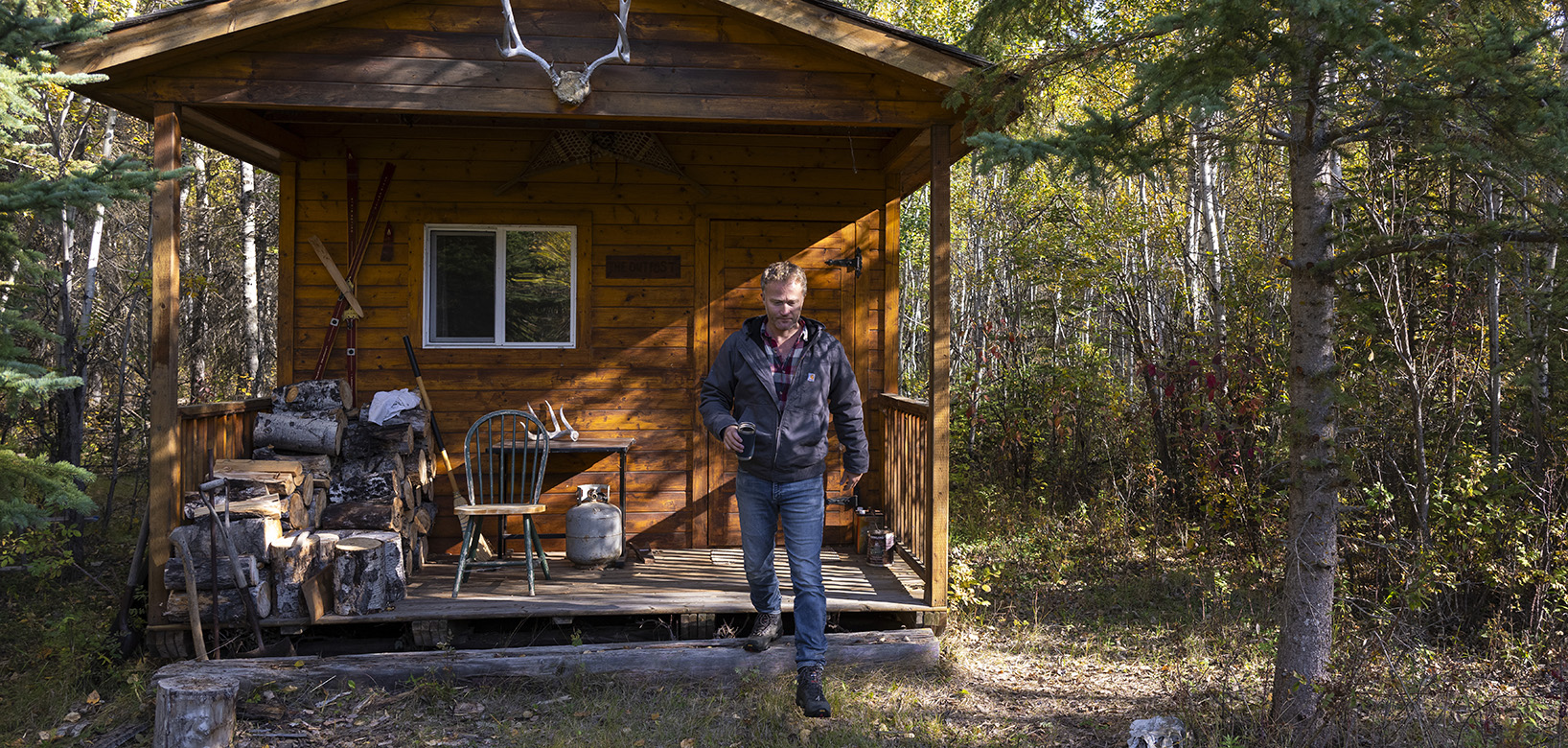 A man with short, light-coloured hair stepping off the front step of a wooden cabin in the middle of the woods.