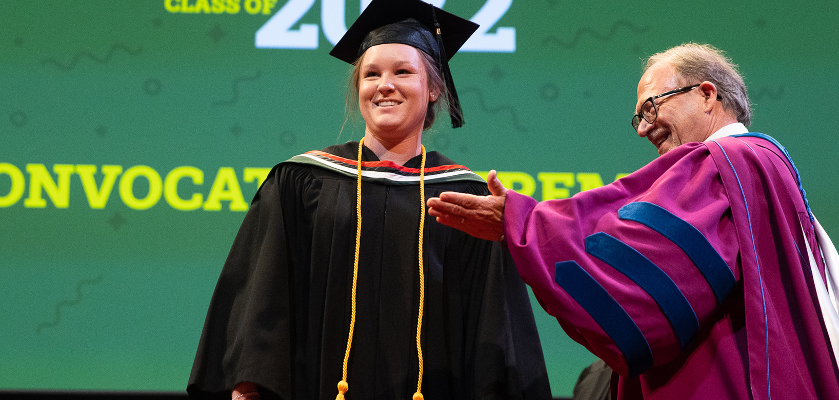 Student in convocation gown and cap smiling on stage in the centre of the shot. Just in front of the student and to the right is the Dean of Augustana in his academic gown, smiling and gesturing towards the student.