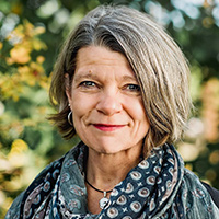 A photo of professor Ardelle Ries, wearing a blue scarf in front of greenery. 