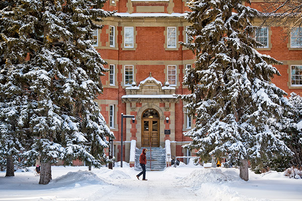 A photo of North Campus' Athabasca Hall building in the snow with a person in a winter coat walking in front of it.