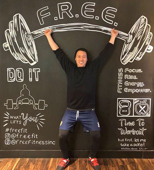 A photo of Tony standing in front of a black chalk board with various images written on it, including a barbell.