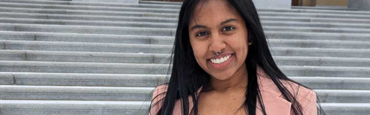 Jewel Anne Naicker standing on stone steps leading up to a tall, stone building. She has dark hair and is wearing a white shirt and pink blazer.