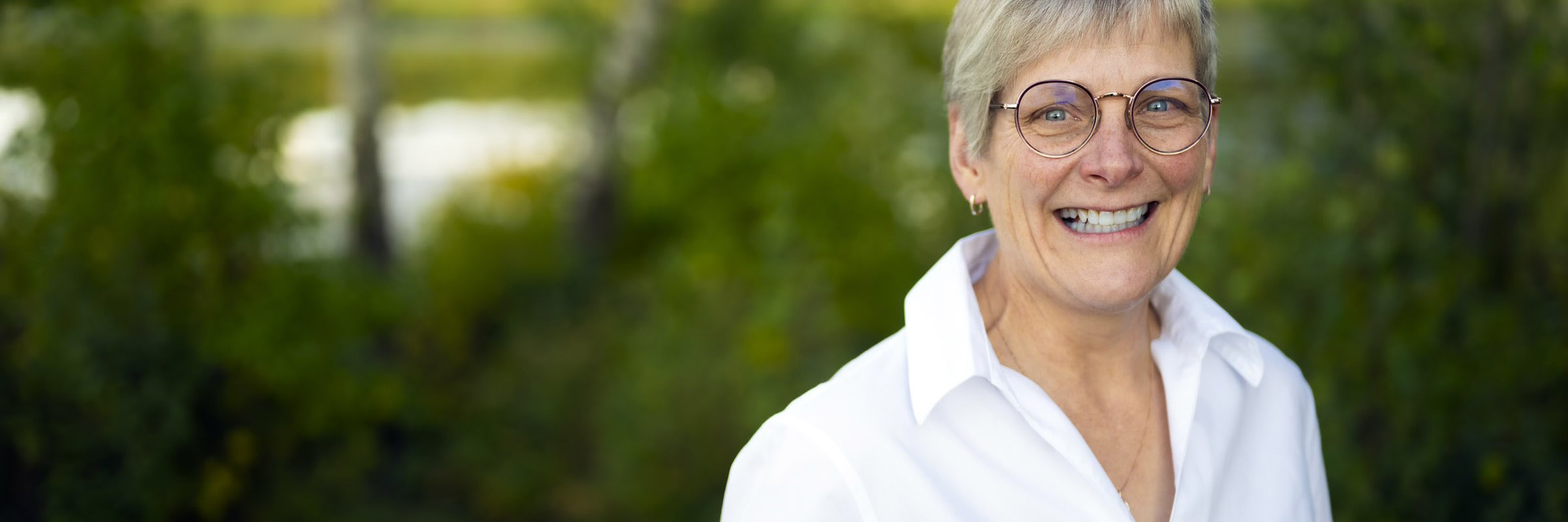 Dee Patriquin smiling at the camera while standing in a forested area.