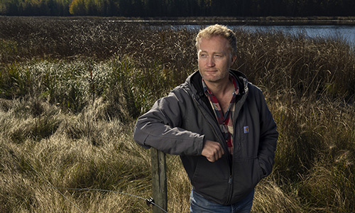 A man with short, light-coloured hair leaning on a fence post in a wetlands area, wearing a plaid button down and zip-up work coat while looking off to the side.