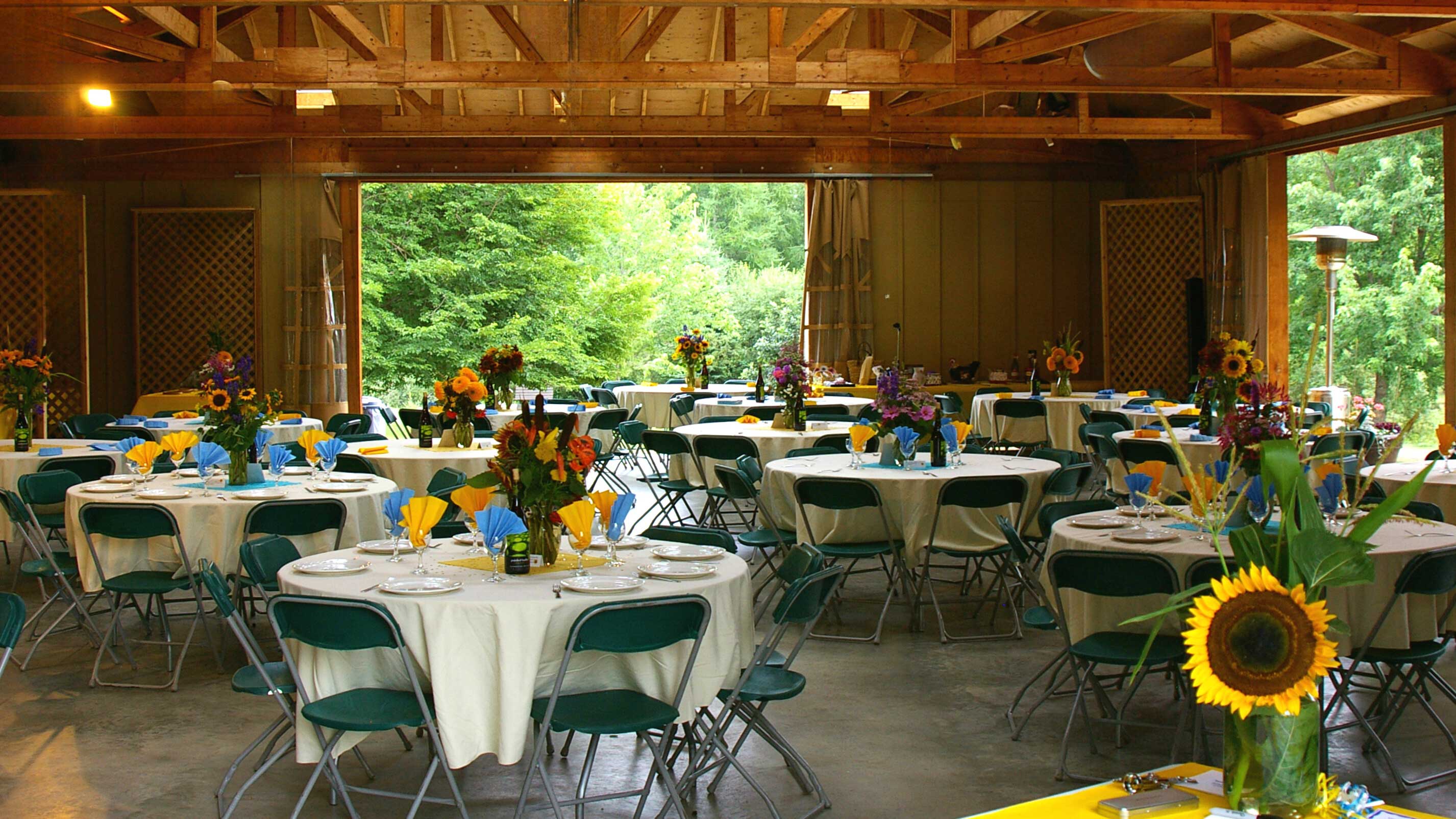 Pine Pavilion setup with round tables with white tablecloths and green chairs. A sunflower in a vase on the lower right hand side