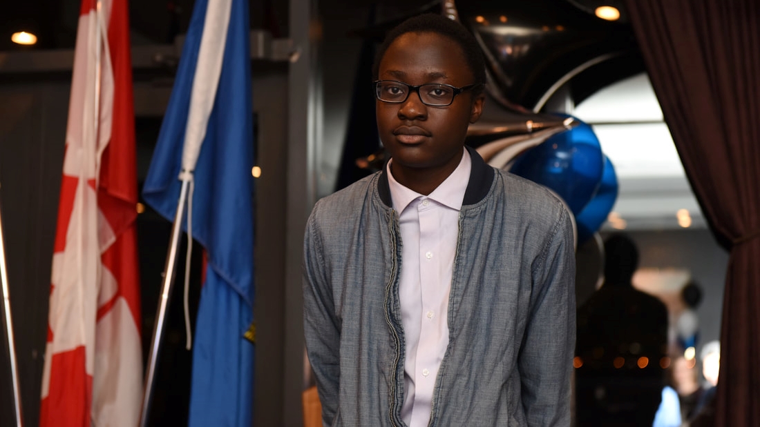 an image of Francis Rweyongeza wearing a denim jacket standing in front of a Canadian and Albertan flag.