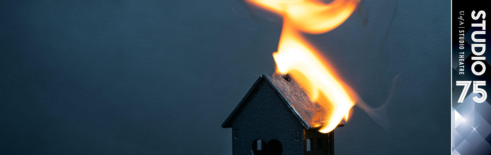 photo of wooden model of a tiny house on a dark background with flames coming off of the roof