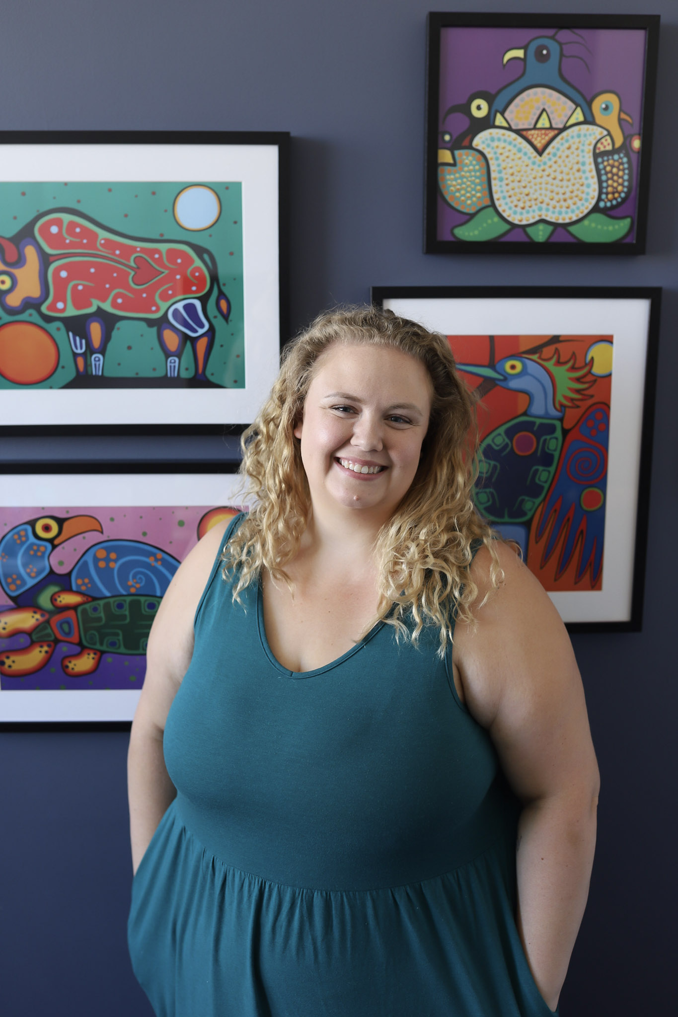 Danielle Gardiner Milln portrait wearing a teal dress with curly hair in front of a wall of paintings