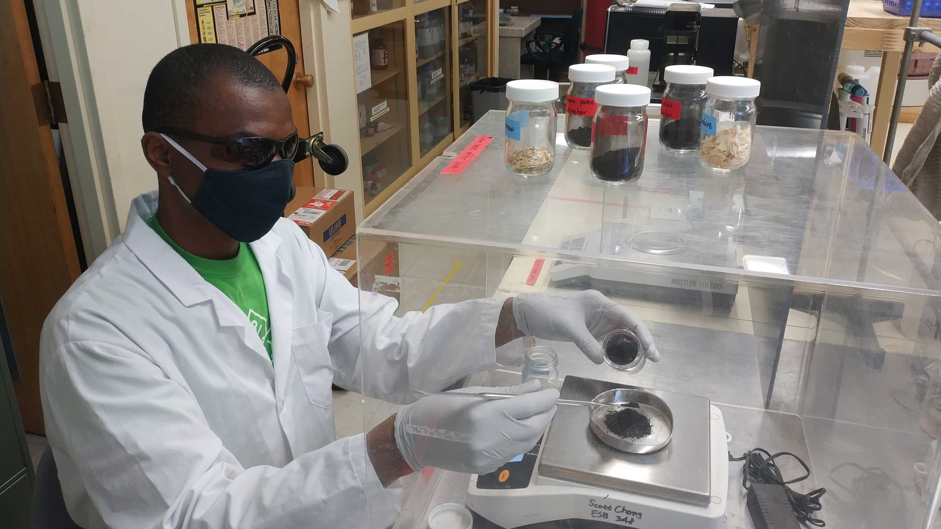 Christopher in white lab coat weighs a sample on a scale at a lab bench