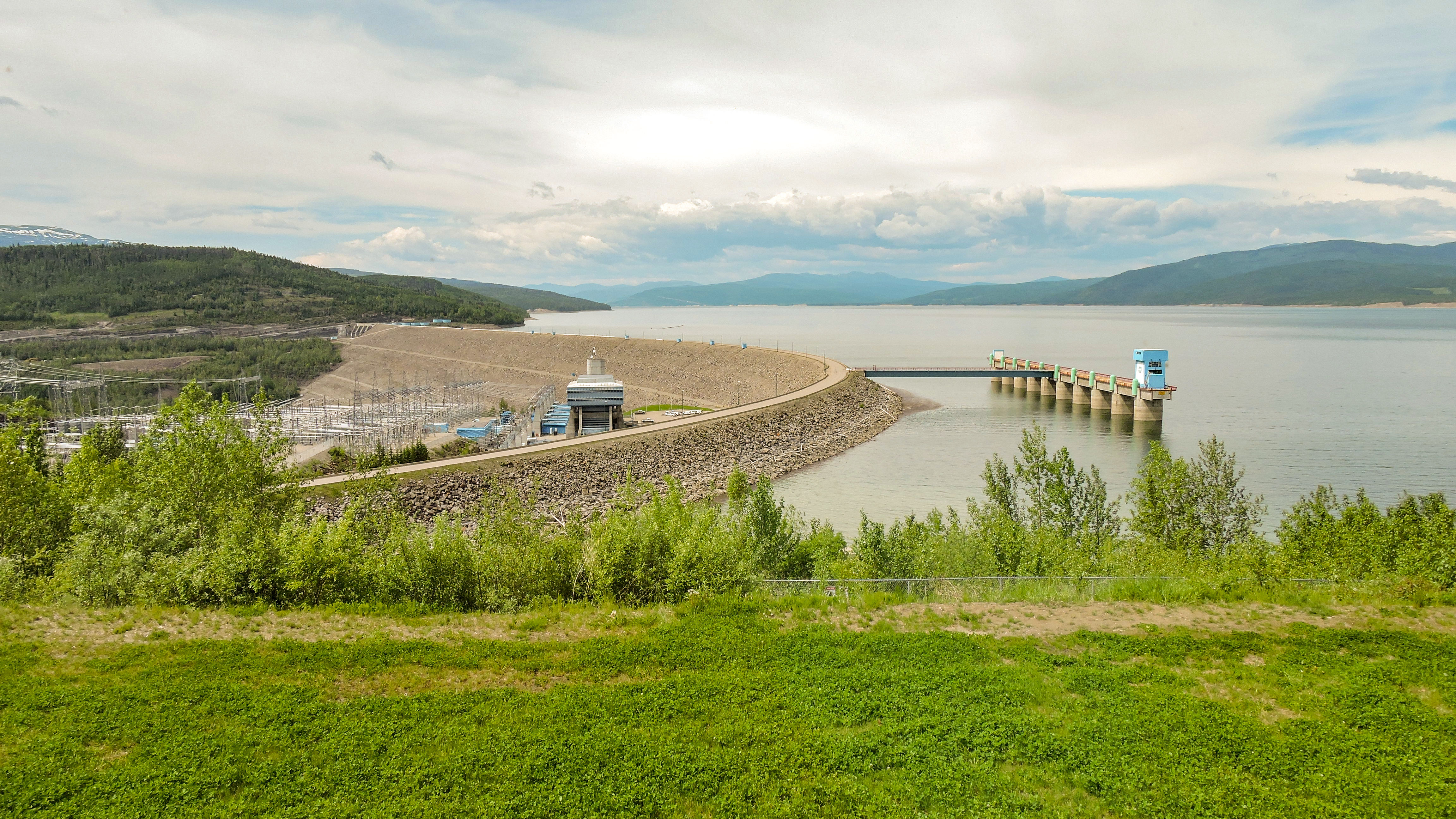 A dam holds back a body of water on the right, with lush grass in front.