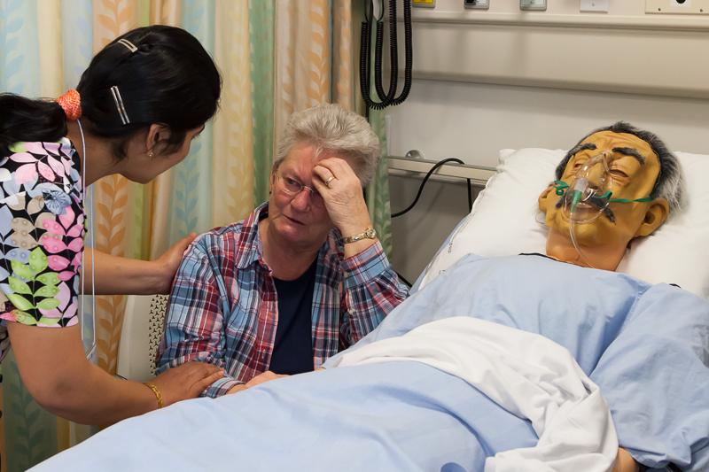 image of patient in hospital bed with a loved one sitting next to them on their left and that loved one is speaking to a medical professional.