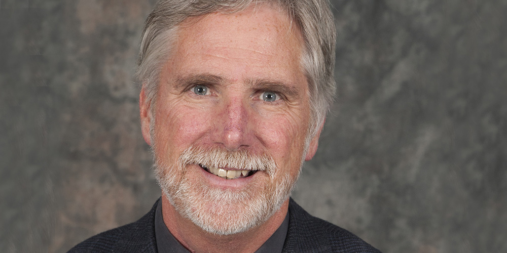 caucasian man with grey hair in black shirt and green tie smiling at camera