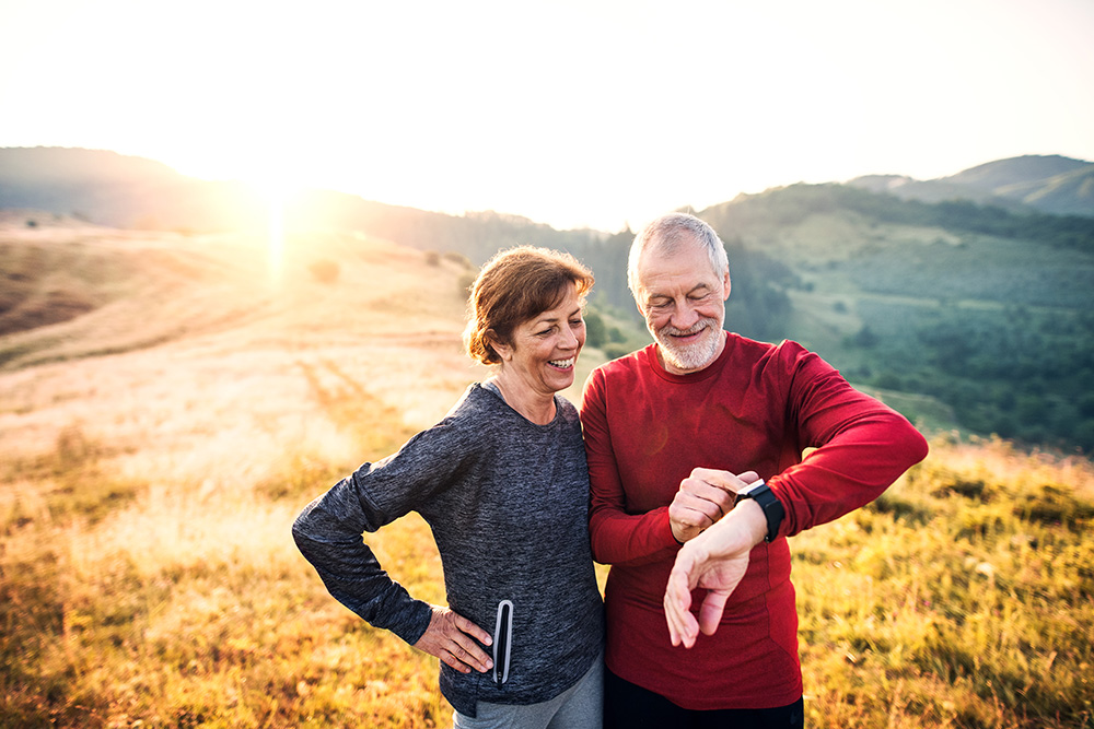 A woman on the left and man on the right both looking at the man's watch. The two are standing in a yellow field.