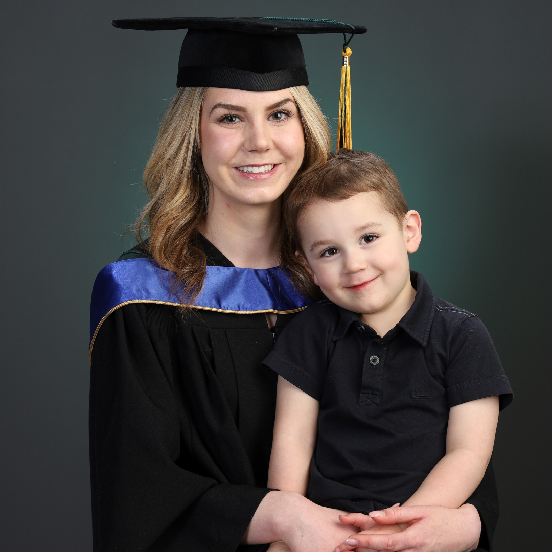 blond woman in cap and gown poses for photo with young son in a black shirt on her lap