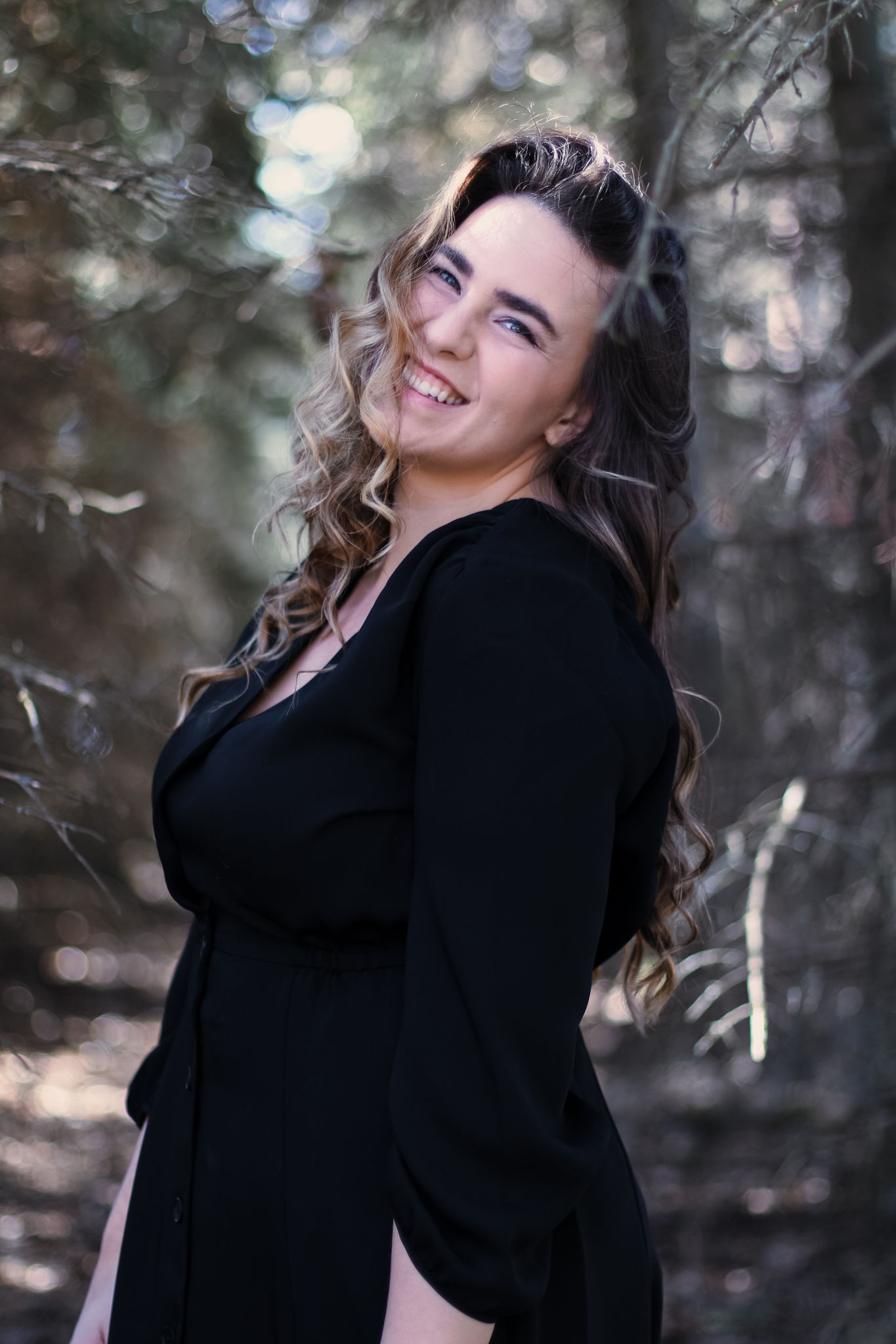 woman in black dress with long brown and blonde hair standing in a tree'd area looking over left shoulder and smiling into camera