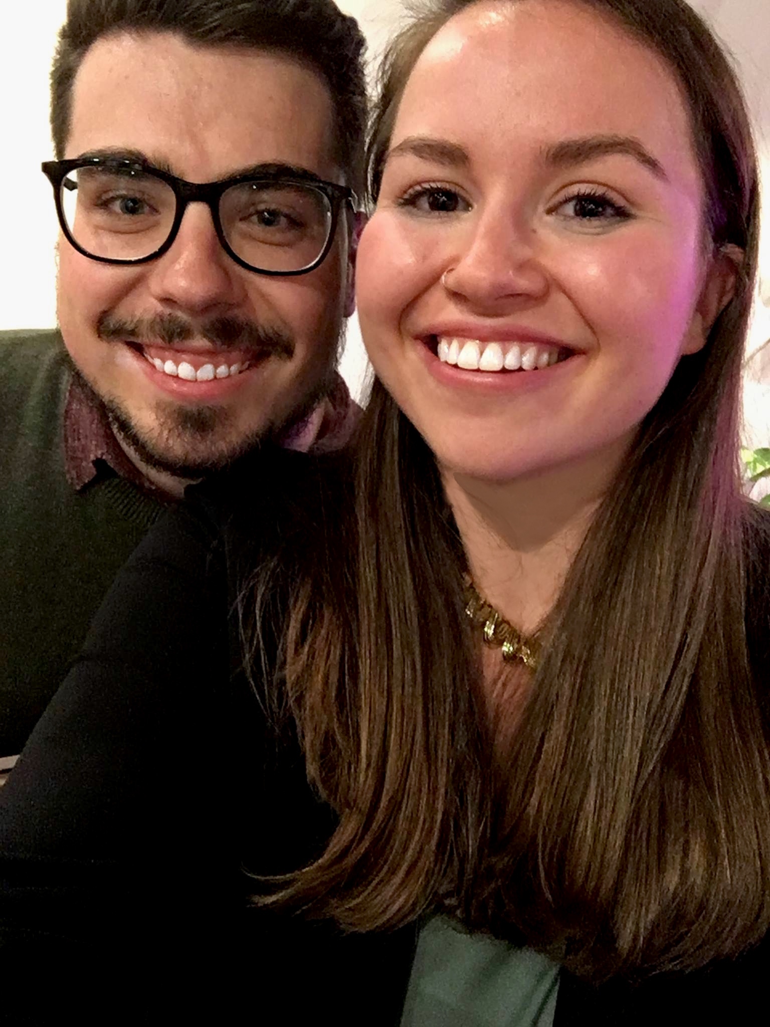 brunette male with glasses and facial hair in green sweater and female with long brunette hair in black sweater smile into camera