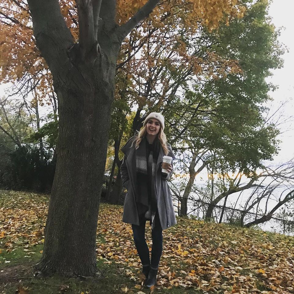 woman with long blonde and red hair wearing a cream-coloured toque drinks coffee in an outdoor treed area during the fall