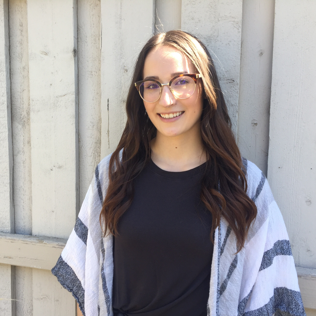 female with long brown hair and glasses wearing a black shirt and black, grey and white shawl standing outside in front of white fence smiles into camera