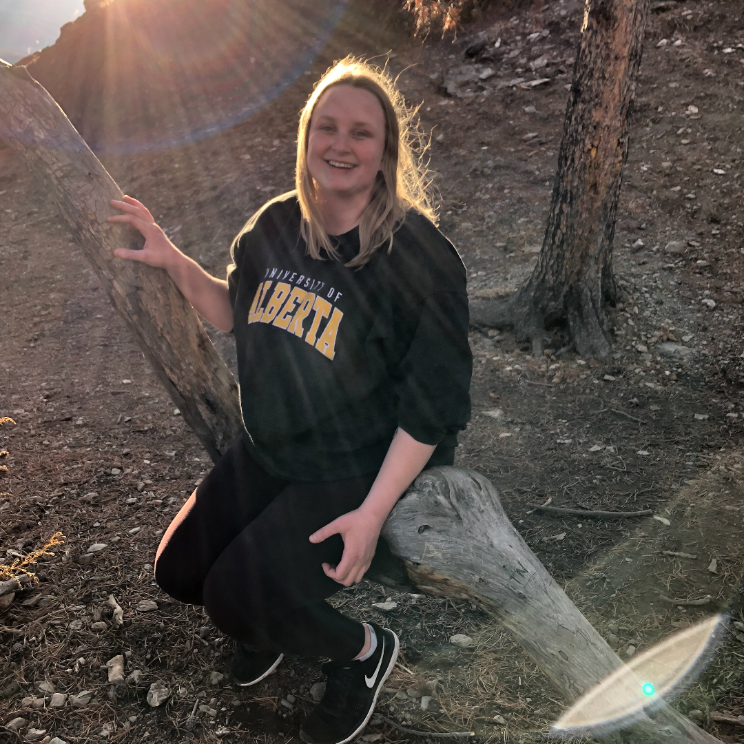 blonde woman wearing a green University of Alberta sweatshirt and black pants sits on tree in wooded area, smiles at camera