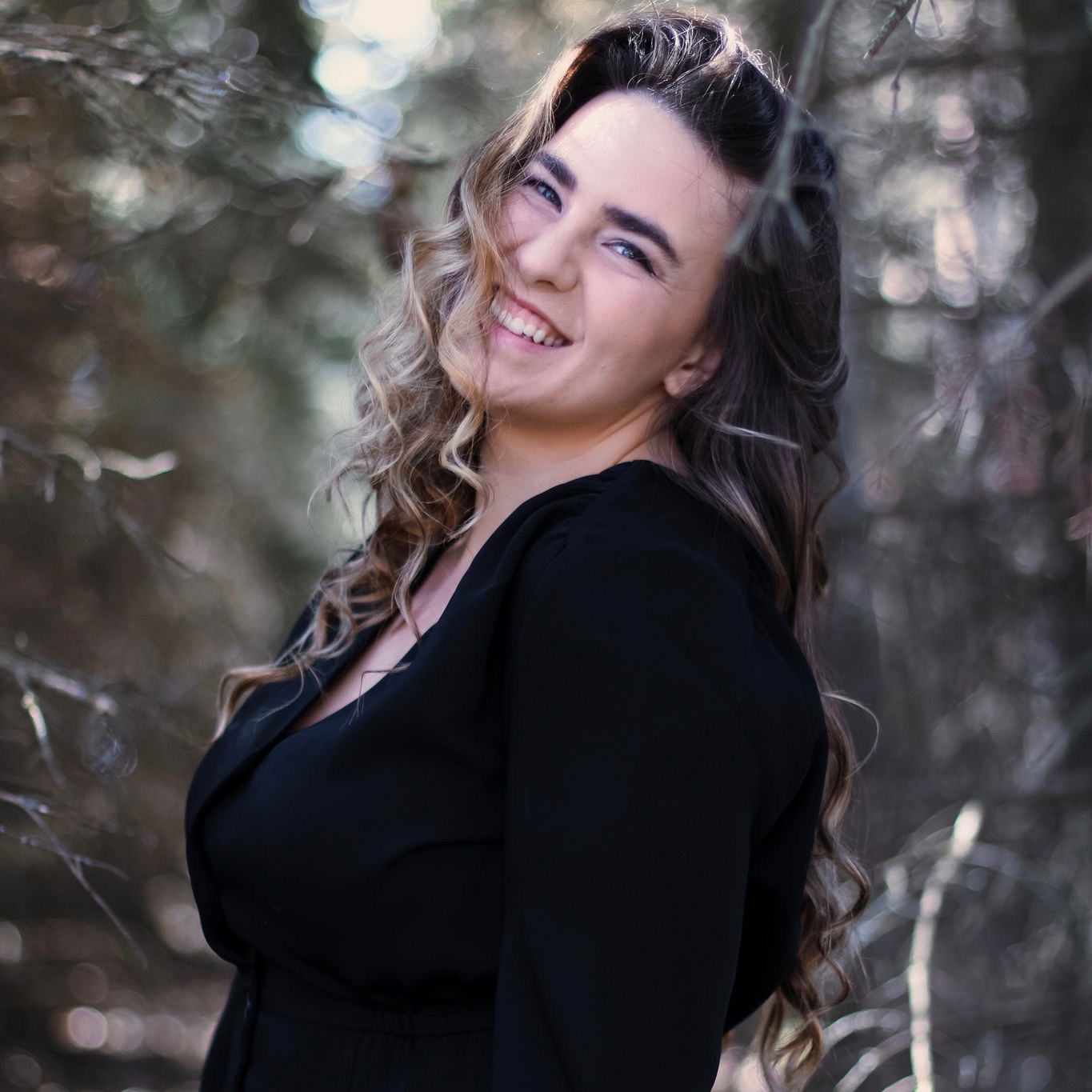 woman in black dress with long brown and blonde hair standing in a tree'd area looking over left shoulder and smiling into camera