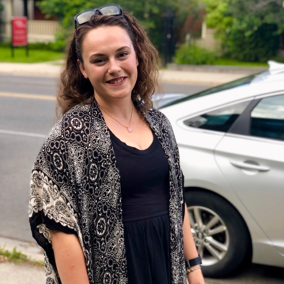 female in a black dress and a black and white komono style cover up smiling at camera while standing outside beside a silver car