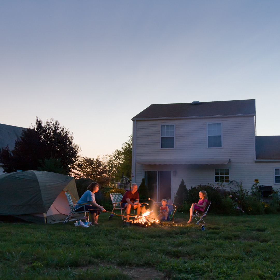 family of four--a mom, a dad, female youth and male toddler--sit around backyard fire roasting marshmallows