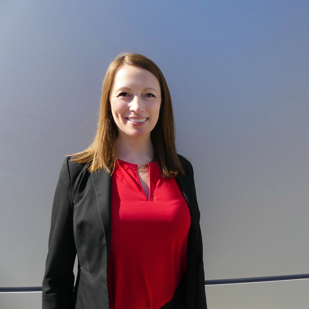 dark haired woman in dark pink shirt and black blazer standing in front of a blue-grey background smiles into camera