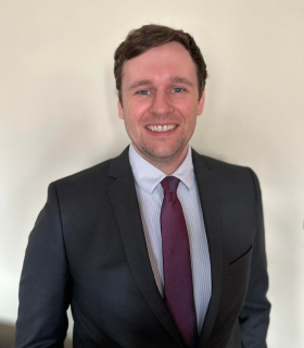 Andrew Sawyers is pictured in a suit and tie against a light backdrop, smiling into the camera