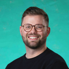 A man with short hair and glasses is picture smiling into the camera against a green studio background