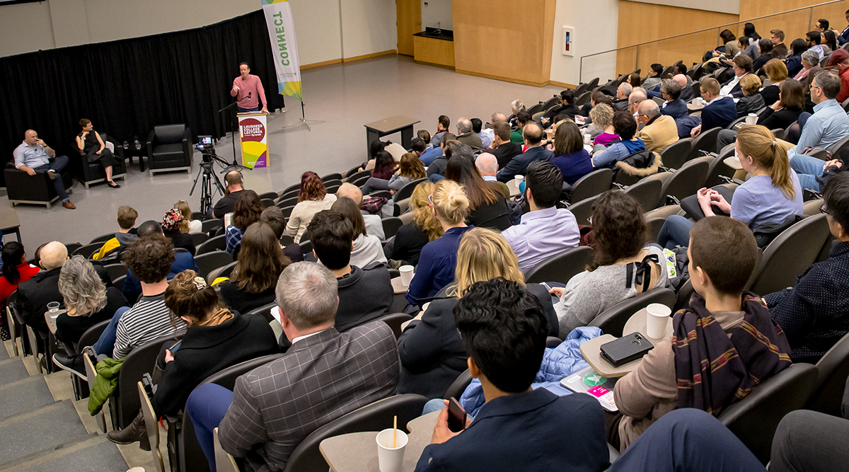 presenters on stage speak in front of a crowd in a lecture theatre