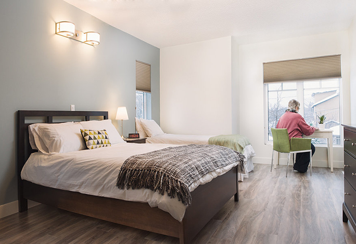 Interior photo of a Compassion House bedroom, which is brightly lit and has a double and single bed, a desk with a woman sitting at it.