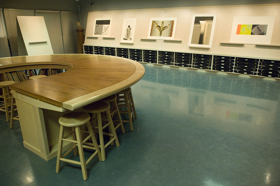 The left side of the Print Study Centre, behind a semi-circle wooden table with a series of books on the surface and several shelving units in the background.