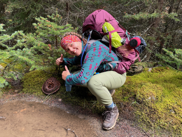 Taylor is crouched down in a forest, smiling at the camera and giving it two thumbs up