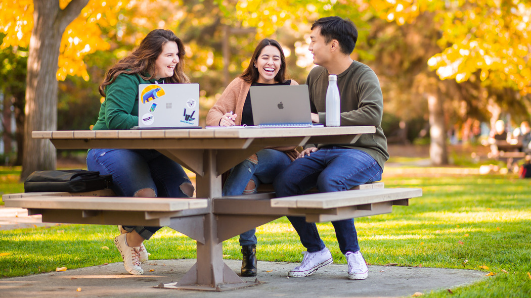 Three students laugh together while sitting on a bench in Main Quad on a sunny fall day.