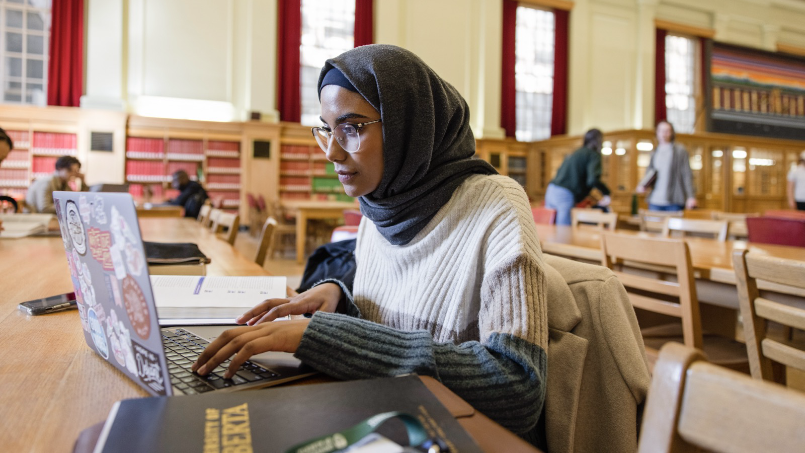 A young woman wearing a hijab sits at a long table in a library, focused on her laptop. She is typing with an open book beside her. The background shows bookshelves and other students studying at similar tables.