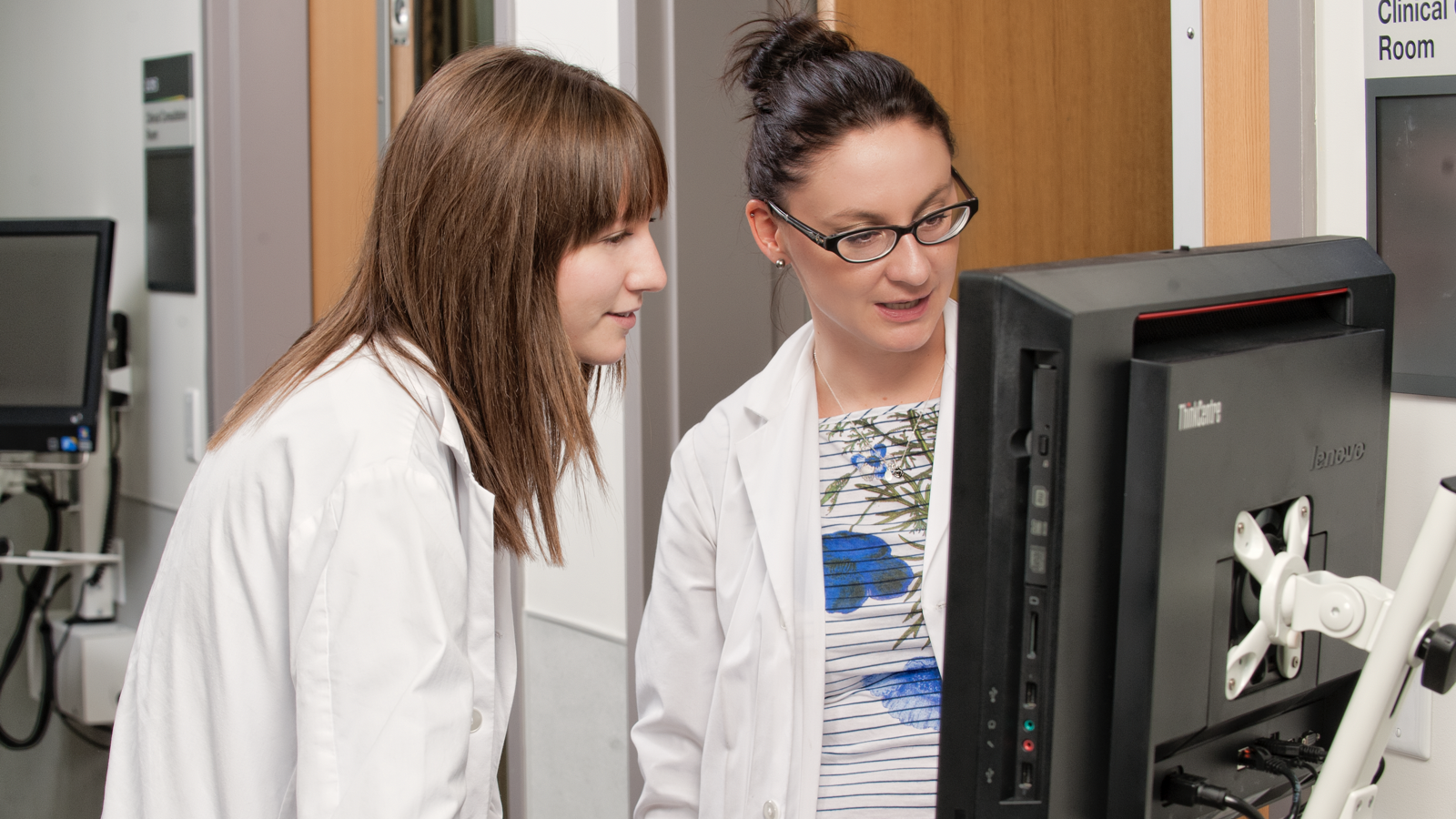 Two researchers looking at a computer screen. They are wearing lab coats and appear to be in a lab.