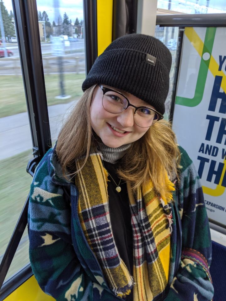  A smiling woman with long blonde hair in a yellow plaid scarf, blue coat, glasses, and black toque